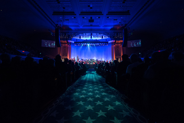 Ein großer Auditorium mit sitzenden Menschen, eine Bühne mit Musikern, die Instrumente spielen, und eine Decke mit Lichtern und Fernsehgeräten, was auf einen Konzertsaal für die Nationale Convention 2019 hindeutet.