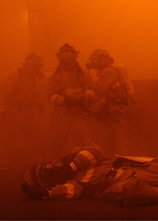Schwarz-weißes Bild eines Feuerwehrmanns, der vor intensiven Flammen auf dem Boden liegt, umgeben von einer Gruppe von Menschen mit Helmen und Sauerstofftanks in der Nähe.