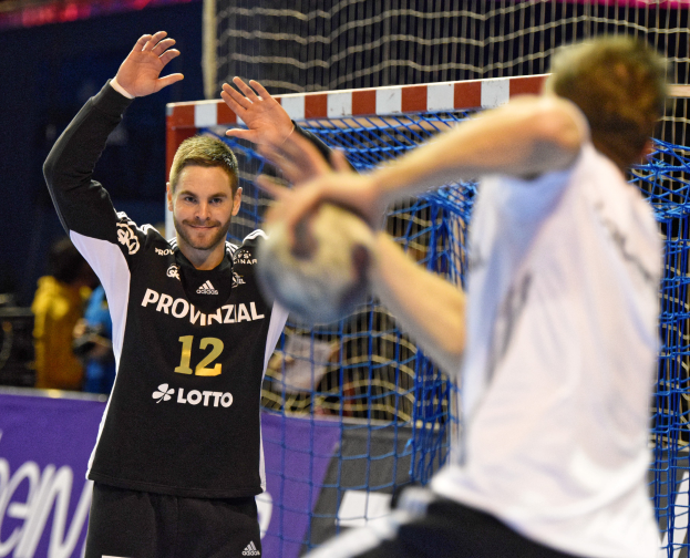 Zwei Männer spielen Handball vor einem Tor mit Netz und einem Banner mit Text im Hintergrund und einigen Zuschauern in einem Stadion.