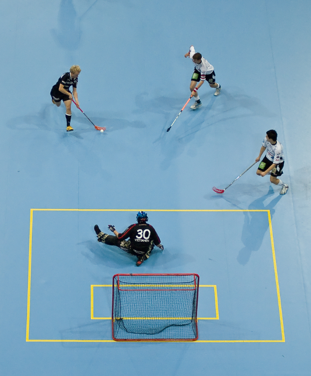 Drei Personen in Sportbekleidung spielen Hockey, wobei eine Person rechts mit einem Hockeystock einen Ball schlägt und eine Person hinter einem Netz auf dem Boden sitzt.