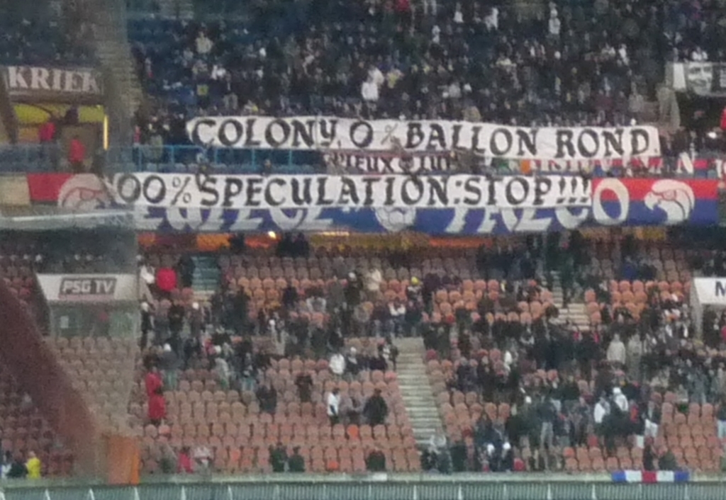 Ein Stadion mit einer großen Menschenmenge, Bannern und Stühlen, mit einem Zaun unten und einer Treppe links.