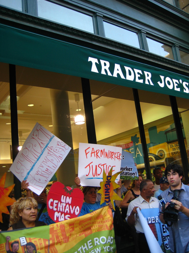 Eine Gruppe von Menschen steht in der Nähe eines Geschäfts und hält Schilder und Plakate in den Händen, wobei hinter ihnen eine Säule und Lichter auf dem Dach zu sehen sind.
