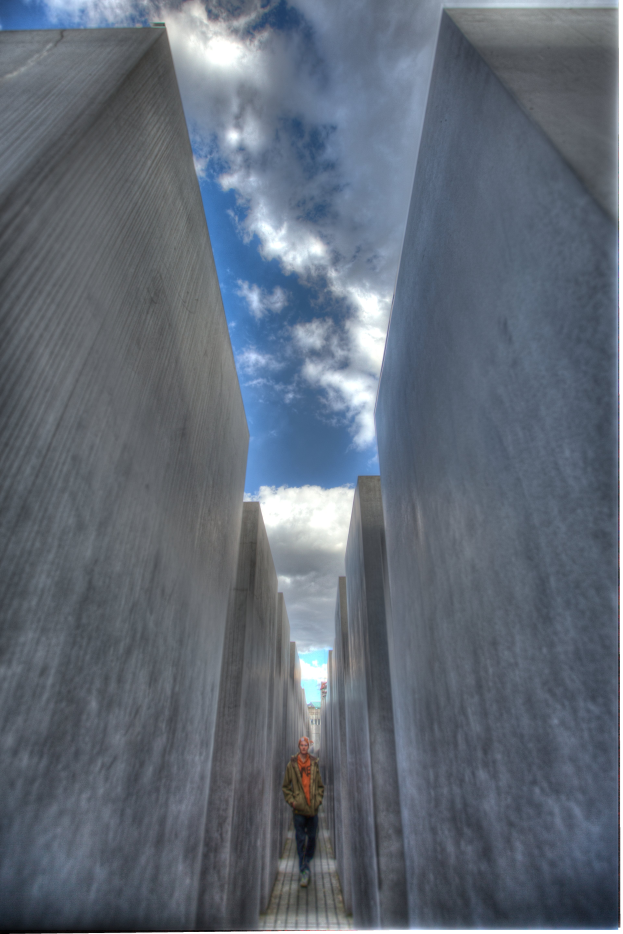 Ein Mann spaziert durch das Holocaust-Mahnmal in Berlin, Deutschland, mit hohen Betonsteinen auf beiden Seiten und einem bew"{o}lktem Himmel im Hintergrund.