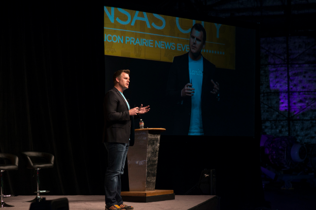 Eine Person in einem schwarzen Hemd spricht auf einer Konferenz, neben einem Tisch mit einer Flasche, mit einem Stuhl dahinter, einem Projektor links von ihnen, der Inhalt anzeigt, und einem schwarzen Vorhang im Hintergrund.