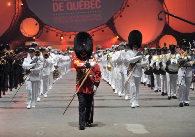 Eine Gruppe uniformierter Personen marschiert durch eine Straße, einige spielen Musikinstrumente, mit einem beleuchteten Schild im Hintergrund, das die Eröffnungszeremonie des Montreal International Festival of Military Bands anzeigt.
