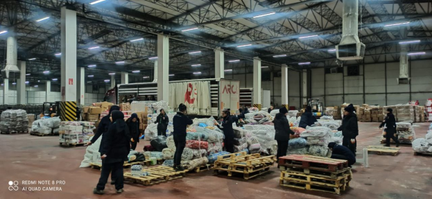 Eine Gruppe von Menschen, die um ein großes Lagerhaus mit Säcken voller Lebensmittel, Holzpaletten, Containern, Pfählen, einem Schild, einer Wand, einem Dach mit Deckenleuchten und Metallpfählen sowie einem Wasserzeichen auf der linken Seite stehen. Es scheint sich um ein Lebensmittelverteilungszentrum in Dubai zu handeln.