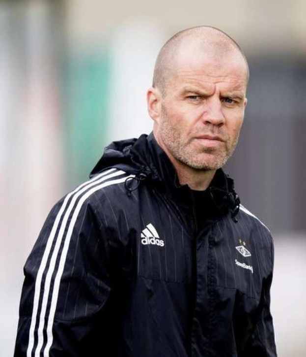 Bald Trainer in marineblauer Jacke mit weißen Streifen und weißem Kragen auf einem Fußballfeld mit gefalteten Händen und ernster Miene.