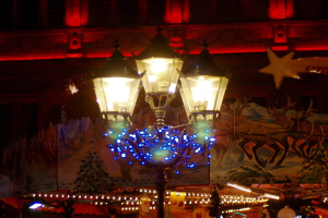Eine mit Lichtern und Festtagsdekoration geschmückte Straßenlaterne steht nachts in einer Stadt, im Hintergrund ist ein Gebäude zu sehen.