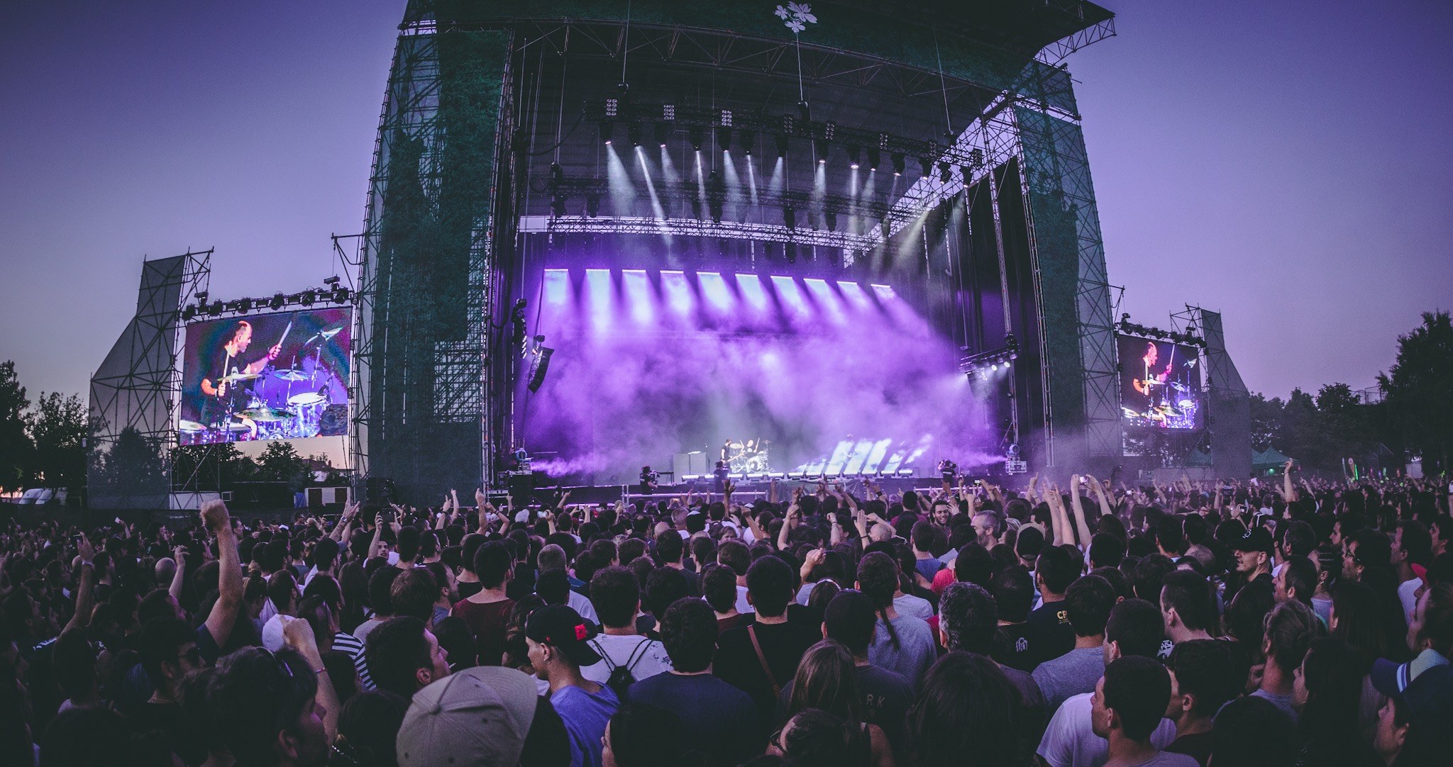 Eine große Menschenmenge auf einem Musikfestival schaut auf der Bühne mit Instrumenten und Bildschirmen, die vor einem Gebäude, Bäumen und einem blauen Himmel aufgebaut sind.