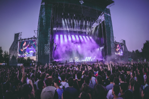 Eine große Menschenmenge auf einem Musikfestival schaut auf der Bühne mit Instrumenten und Bildschirmen, die vor einem Gebäude, Bäumen und einem blauen Himmel aufgebaut sind.