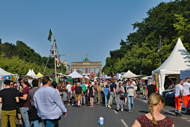 Menschenmenge auf einer Straße mit Zelten, Fahrzeugen und Bäumen an einem Event mit Fahnenmasten, einem Bogen und einem klaren blauen Himmel im Hintergrund.