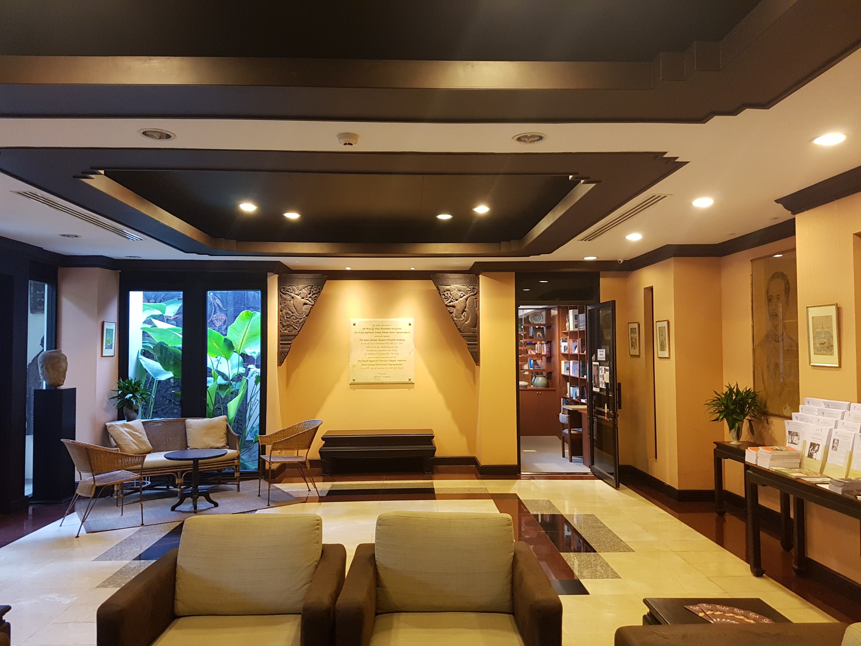 Eine geräumige Hotellobby mit Sitzbereichen, Dekorationspflanzen, Bücherregalen, Wandkunst und Dekoration auf Beistelltischen, beleuchtet von Deckenlampen und natürlichem Sonnenlicht durch Fenster und Türen.