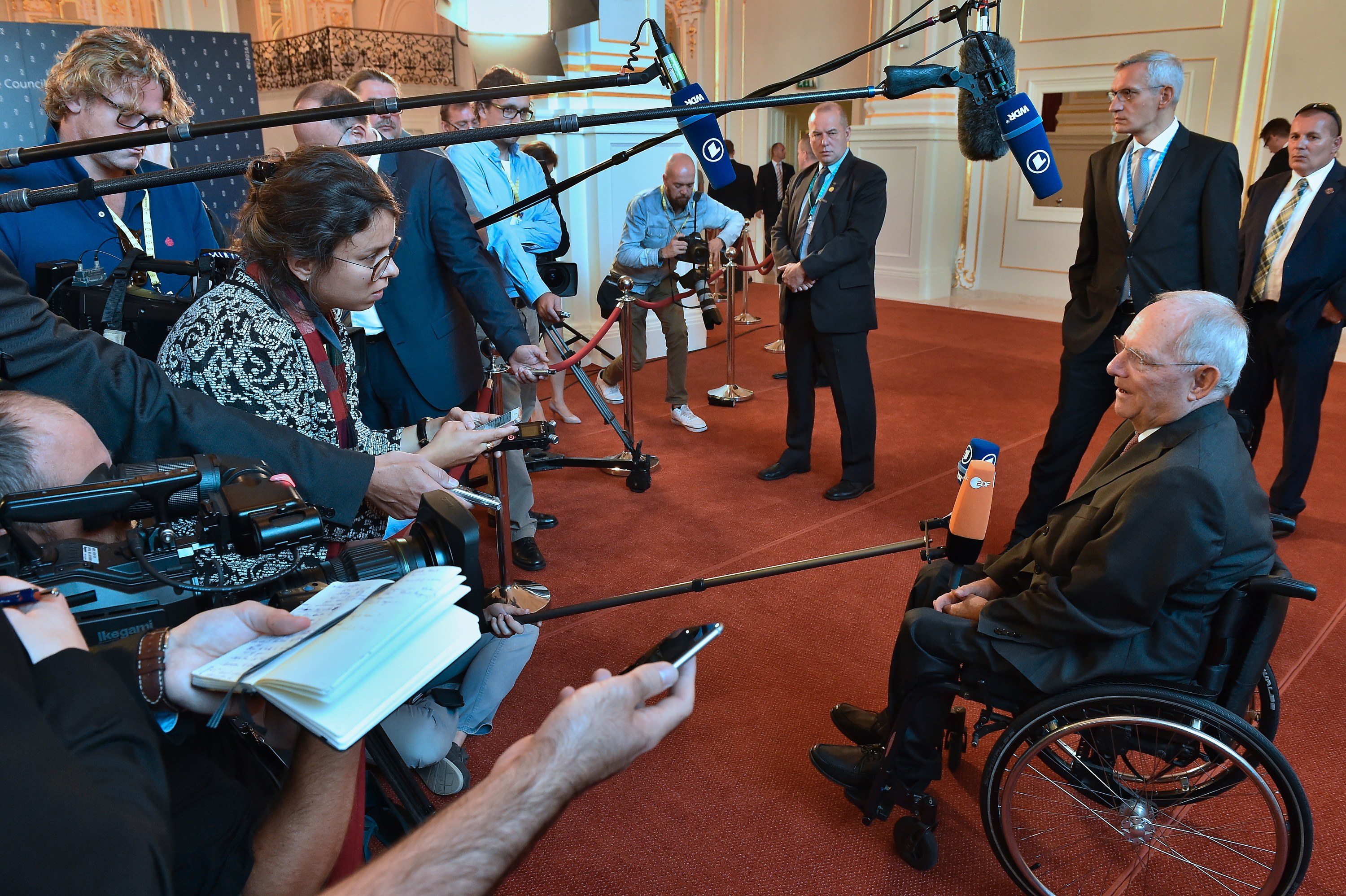Ein Mann im Rollstuhl in Anzug und Krawatte von Reportern mit Kameras und Mikrofonen umgeben, mit einem Banner und einer Tür im Hintergrund.