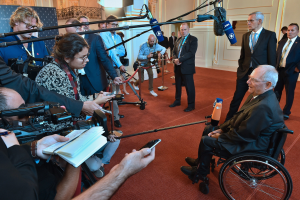 Ein Mann im Rollstuhl in Anzug und Krawatte von Reportern mit Kameras und Mikrofonen umgeben, mit einem Banner und einer Tür im Hintergrund.