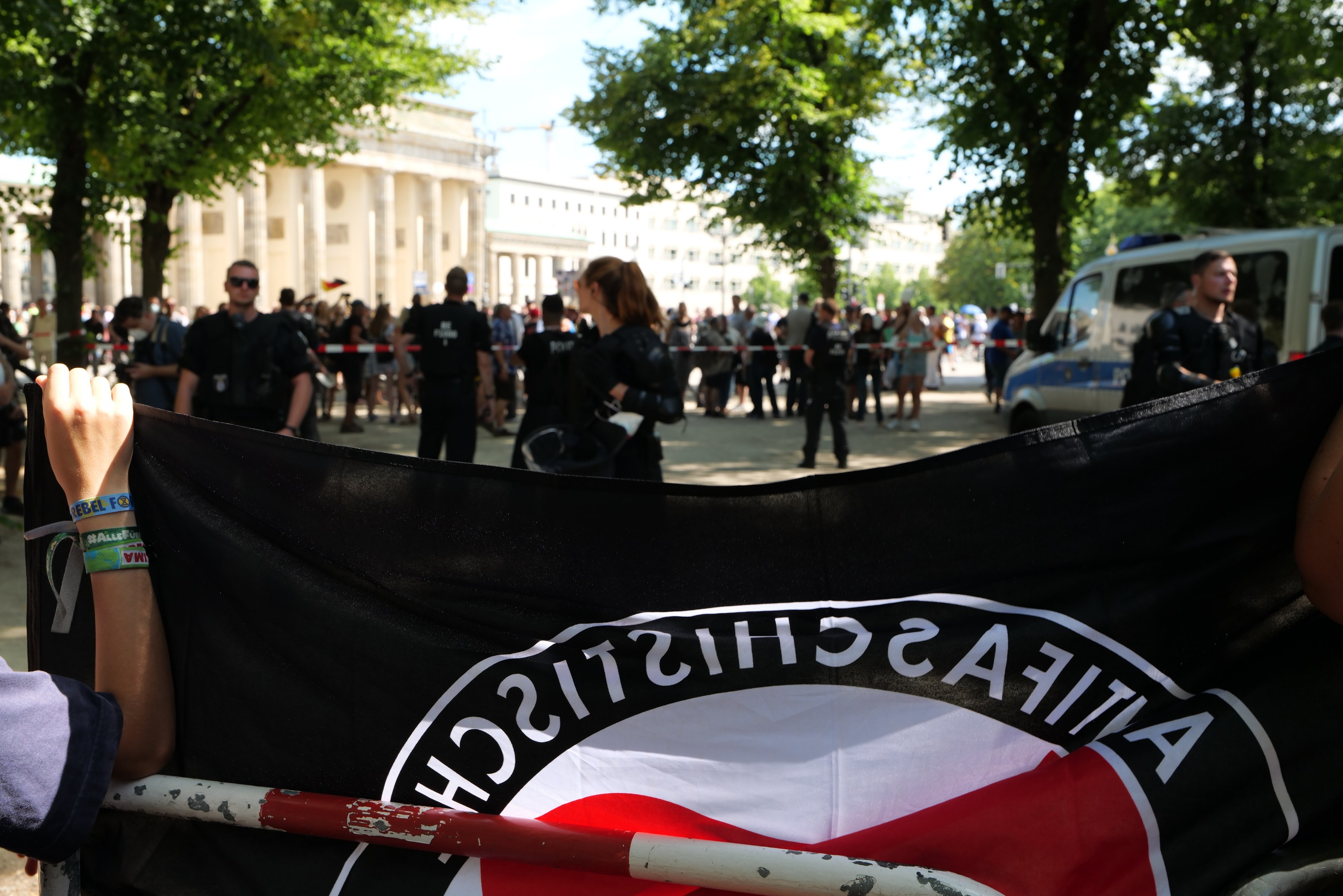 Gruppe von Menschen auf dem Boden, einige halten eine deutsche Flagge vor einer Menge, mit einem Metallpfahl, Fahrzeugen auf der Straße, einem Zaun, Bäumen, Gebäuden mit Fenstern und einem bewölkten Himmel.