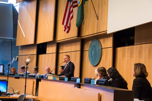 Eine Gruppe von Menschen sitzt an einem Tisch mit Namensschildern, Mikrofonen und anderen Gegenständen, mit einem Mann in der Mitte, vor einem Bildschirm, mit Flaggen und einer Tafel im Hintergrund, was auf eine Ratssitzung hindeutet.