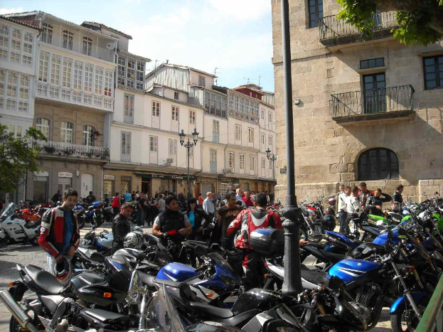 Ein cremefarbenes Gebäude mit zahlreichen Fenstern, das an ein Backsteingebäude mit einer Metall-Fensterfront angrenzt, mit mehreren geparkten Fahrrädern und Menschen, die davor diskutieren.