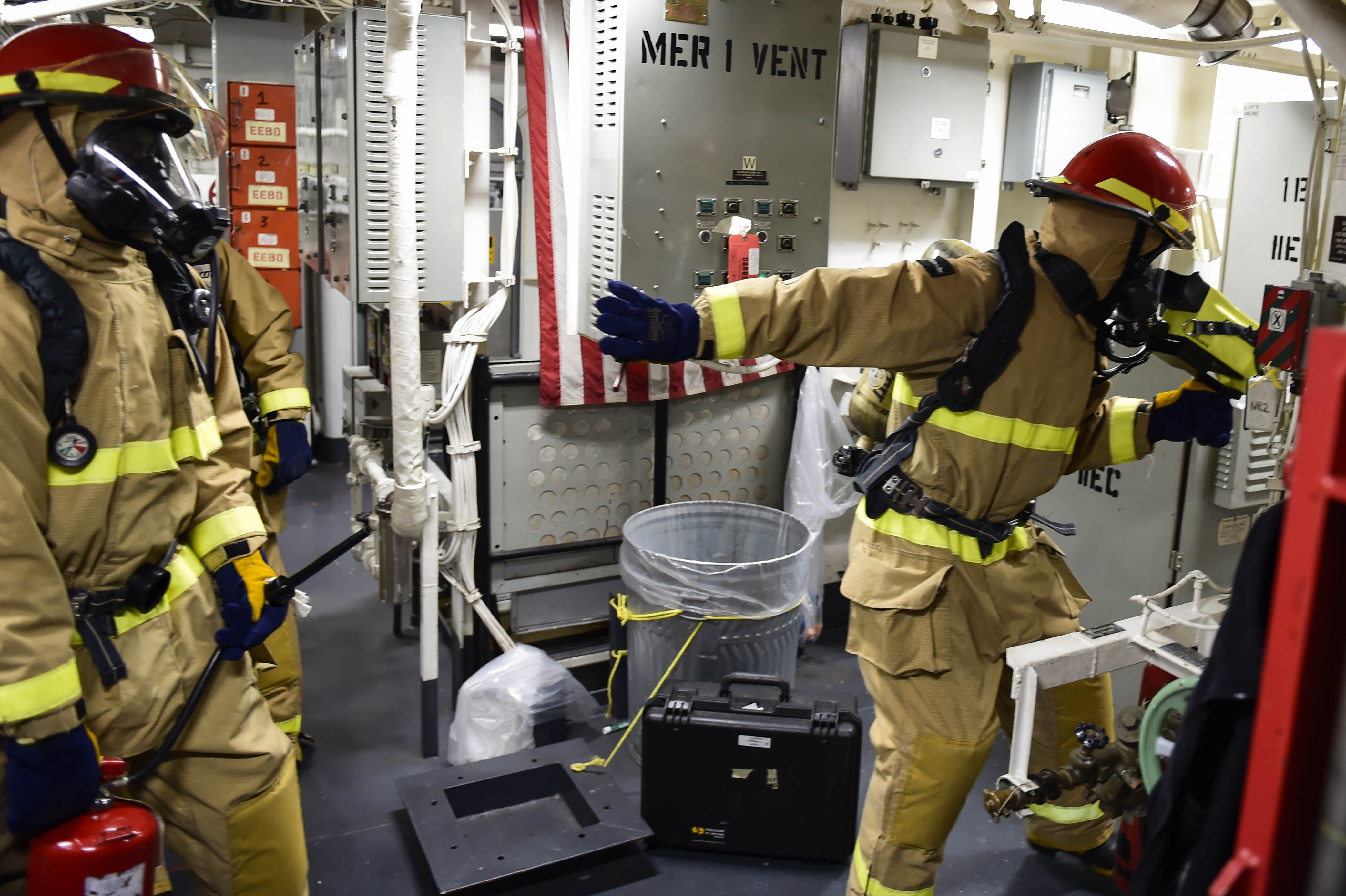 Drei Feuerwehrleute in Schutzausrüstung stehen zusammen in einem Raum mit einem Feuerlöscher links, verschiedenen Gegenständen auf dem Boden, einer Maschine rechts und Rohren und Lampen oben.