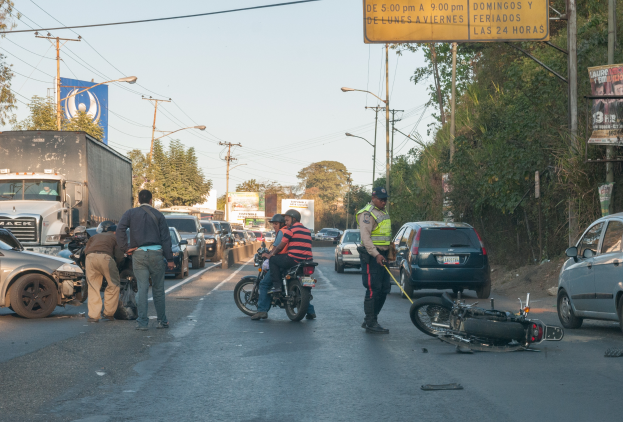 Eine Gruppe von Menschen umringt ein verunglücktes Motorrad am Straßenrand mit mehreren Fahrzeugen, darunter ein Lastwagen, und einem Hintergrund aus Bäumen, Pfählen, Lampen und Schildern unter dem Himmel.