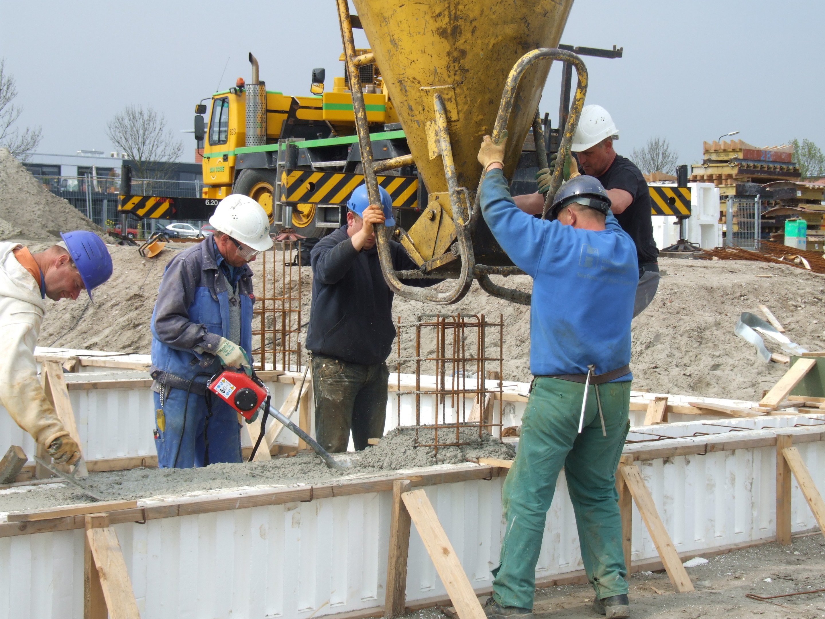 Gruppe von Männern mit Helmen, die eine Betonmischmaschine auf einer Baustelle bedienen, mit Fahrzeugen, Bäumen, Gebäuden und einem klaren blauen Himmel im Hintergrund.
