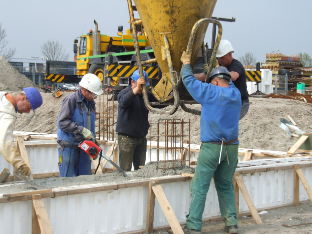Gruppe von Männern mit Helmen, die eine Betonmischmaschine auf einer Baustelle bedienen, mit Fahrzeugen, Bäumen, Gebäuden und einem klaren blauen Himmel im Hintergrund.