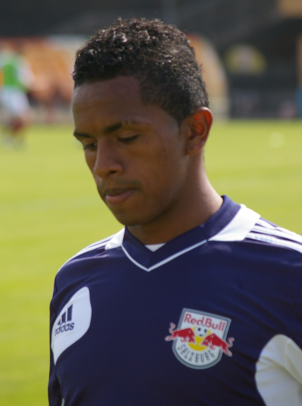 Ein junger Mann in einem blauen T-Shirt steht auf einem Fußballfeld, der als Flügelspieler mit kurzem, dunklem Haar und entschlossener Miene erscheint, vor einem unscharfen Hintergrund.