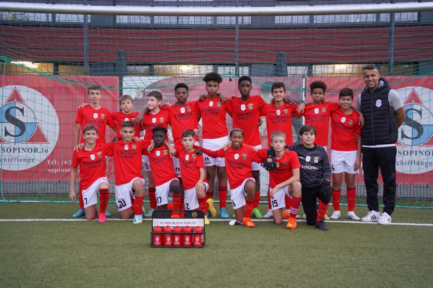 Eine Gruppe junger Männer in roten T-Shirts und schwarzen Shorts steht auf einem Fußballfeld vor einer Kiste roter Gegenstände, im Hintergrund Schilder mit der Aufschrift "FC Rot Schwarz Eglaute - Champions League".