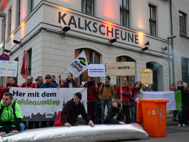 Eine Gruppe von Menschen steht vor einem Gebäude mit Schildern und Plakaten, zwei Personen sitzen im Vordergrund und ein Müllcontainer rechts, bei einer Demonstration in Deutschland.