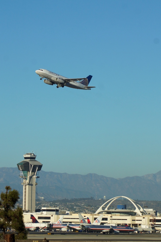 Ein Flughafenblick mit einer weißen Kontrollturm im Vordergrund, parkenden Flugzeugen am Boden, entfernten Gebäuden und einem Flugzeug am Himmel.