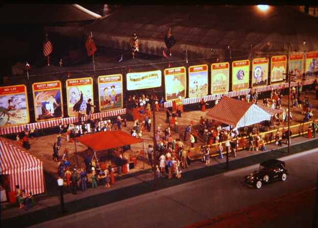 Ein altes nächtliches Karnevalsbild mit Menschen, einem Auto, Polen mit Fahnen, einem Zaun, einem Zelt, Brettern mit Bildern und Text sowie einer Straßenlaterne.
