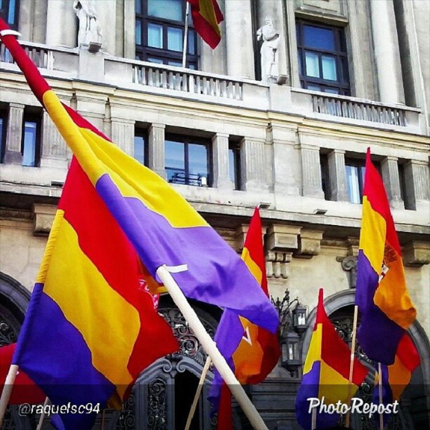 Ein mehrstöckiges Gebäude mit zahlreichen Fenstern und Türen, mit mehreren Pfählen davor, an denen Flaggen in Rot, Gelb und Lila wehen.