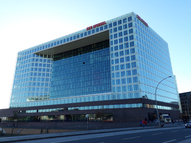 Ein großes Gebäude mit vielen Fenstern, identifiziert als Hauptsitz der Deutschen Bank, umgeben von Straßenlaternen, Autos, Bäumen und einem bewölkten Himmel.