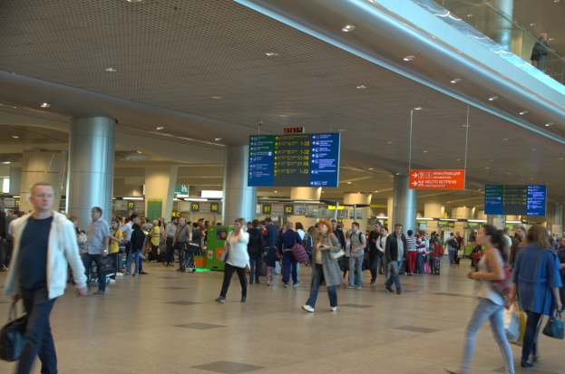 Menschen gehen durch einen Flughafen mit Hinweistafeln, Pfeilern, Deckenleuchten und einer Glasbrüstung auf der rechten Seite.