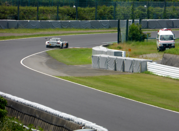 Ein Rennwagen fährt auf einer Rennstrecke mit einem Polizeiwagen im Hintergrund, umgeben von Gras, Pflanzen, Bäumen und Zäunung, mit einem Fahrzeug auf der rechten Seite und Text unten.