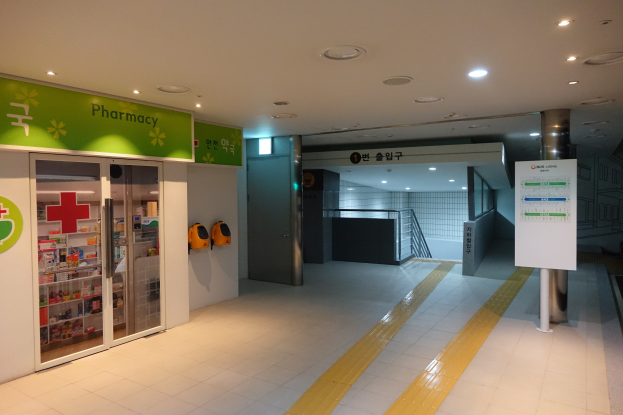 Apotheken-Eingang mit einem "Apotheke"-Schild auf der linken Seite, einer Tafel mit Text auf der rechten Seite, Hintergrundobjekte und beleuchtete Dachlichter.