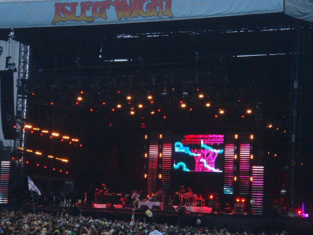 Eine B√ľhne mit Lichtern, einem Bildschirm und Musikern, die Instrumente spielen, mit einer Fahne √∂ber ihnen, einer Menge unten und Flaggen mit St√§ben und Ballons drumherum.