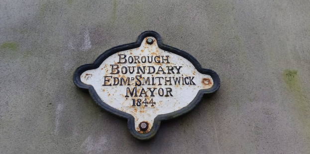 Metallplatte an der Außenseite eines Gebäudes, die die Borough-Grenze markiert, mit schwarzem Hintergrund und weißer, fetter, zentrierter Schrift, die "Borough Boundary Edm Smithwick Mayor" lautet.