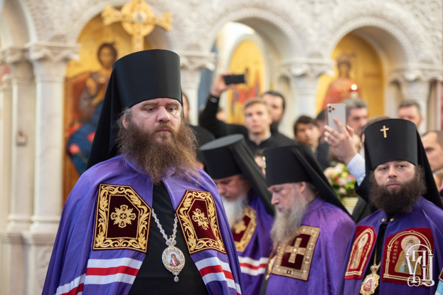 Eine Gruppe von Priestern, die in einer Kirche nebeneinander stehen, mit einer Person im Vordergrund, die ein Mobiltelefon hält, Säulen und ein gerahmtes Foto im Hintergrund.