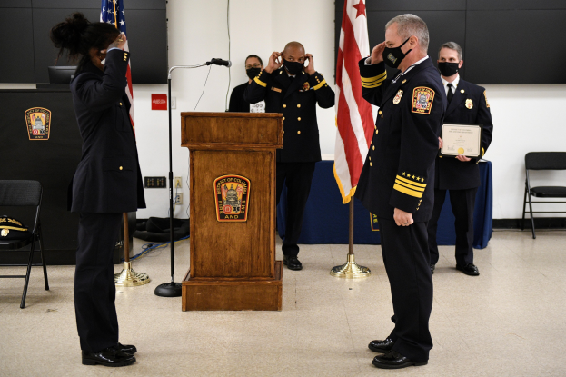Gruppe uniformierter Personen in Masken vor einem Rednerpult mit Mikrofon, die eine Urkunde während einer Vereidigung eines neuen Feuerwehrchefs halten, umgeben von Fahnen, Stühlen, Tischen und einer Wand.