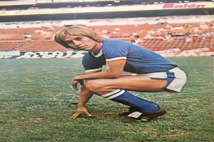 Ein Mann kniet auf einem Fußballfeld umgeben von Menschen, die auf Stühlen und Tafeln mit Text sitzen.