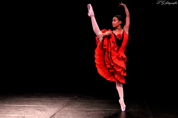 Eine Frau in einem roten Kleid tanzt anmutig auf der Bühne, ihre Arme und Beine ausgestreckt, trägt Pointe-Schuhe und hat ihr Haar zu einem ordentlichen Knoten gebunden.