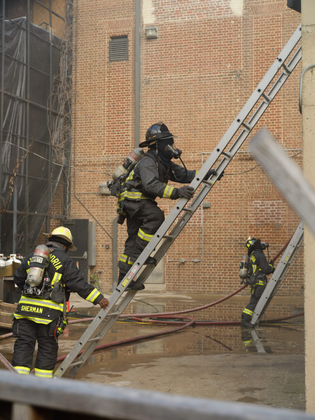 Feuerwehrleute in Helmen und Lufttanks klettern an einer Leiter vor einem Backsteingebäude mit Rohren auf dem Boden und einer Metallstange unten, während im Hintergrund ein weiteres Gebäude mit Fenstern und Netz zu sehen ist.
