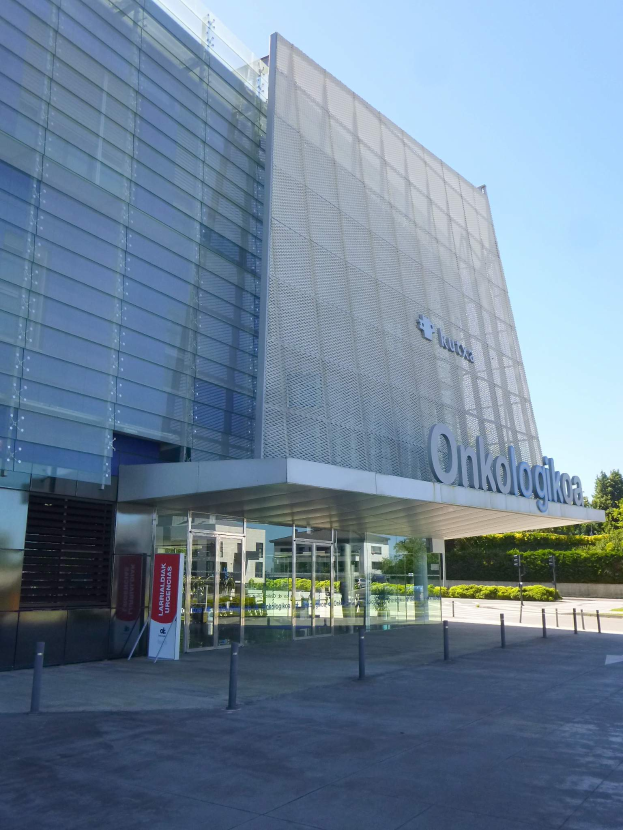 Ein großes Gebäude mit zahlreichen Fenstern, umgeben von Pfählen, ein Schild mit der Aufschrift "Onkologika, das größte medizinische Zentrum der Welt", eine Gruppe von Bäumen, Pflanzen und ein bewölkter Himmel.