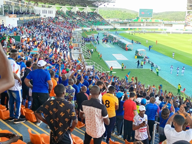 Eine große Menschenmenge versammelt sich vor einem eingezäunten Fußballfeld, wobei einige auf Stühlen sitzen und andere stehen, mit Blick auf Hügel unter einem klaren blauen Himmel während der Afrikameisterschaft 2019.