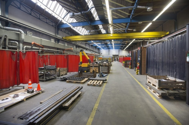 Großes Lagerinnere mit Holzpaletten, Metallobjekten, Verkehrskegeln und Maschinenfahrzeug, mit roten Vorhängen, Deckenlampen und Rohren.