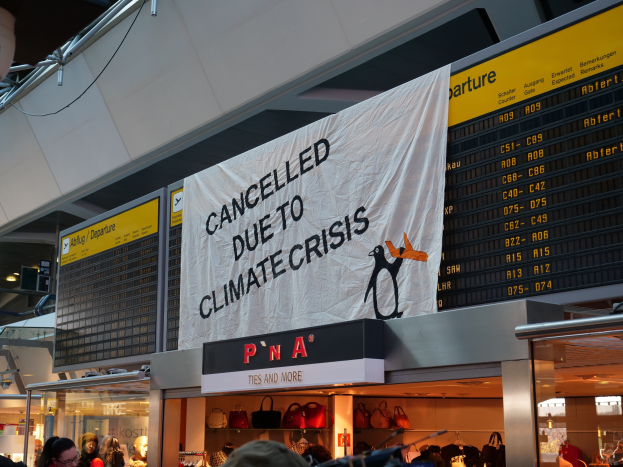Gruppe von Menschen vor einer Flughafen-Abflugtafel mit einem Banner "Aufgrund der Klimakrise gestrichen", Gepäckwagen sichtbar und beleuchtete Deckenlampen