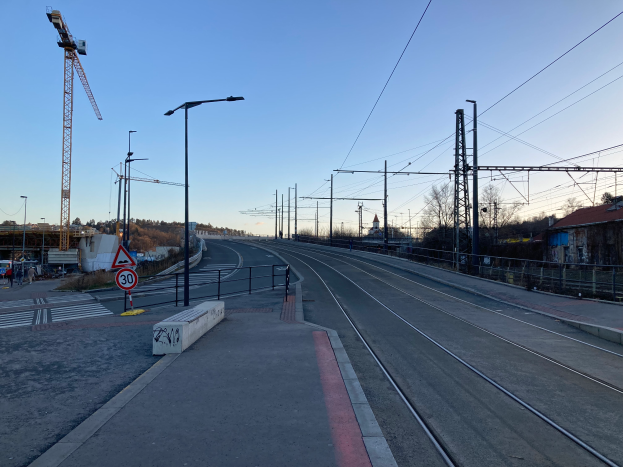 Eine Bahnstrecke mit einer Baustelle im Hintergrund, Strommasten mit Drähten auf der rechten Seite, eine Straße mit Laternenmasten, Schildern, Geländern und einigen Menschen auf der linken Seite, eine Brücke in der Mitte, Bäume und Gebäude vor der Brücke, der Himmel oben und Text unten.
