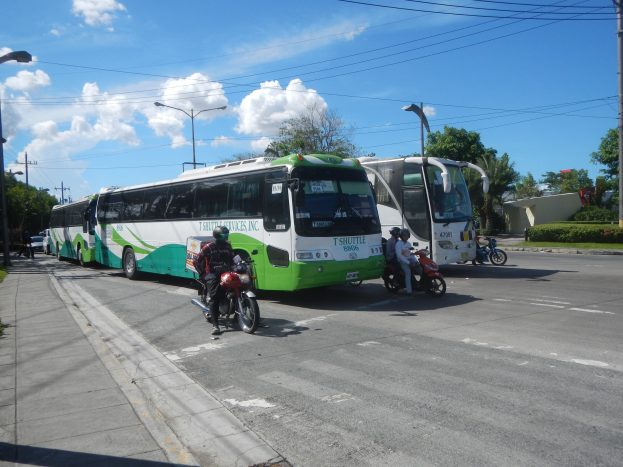 Ein grüner und weißer Shuttlebus steht am Straßenrand mit Motorrädern davor, ein grasbewachsener Fußweg links daneben und Gebäude, Bäume und Laternen im Hintergrund unter einem klaren blauen Himmel.
