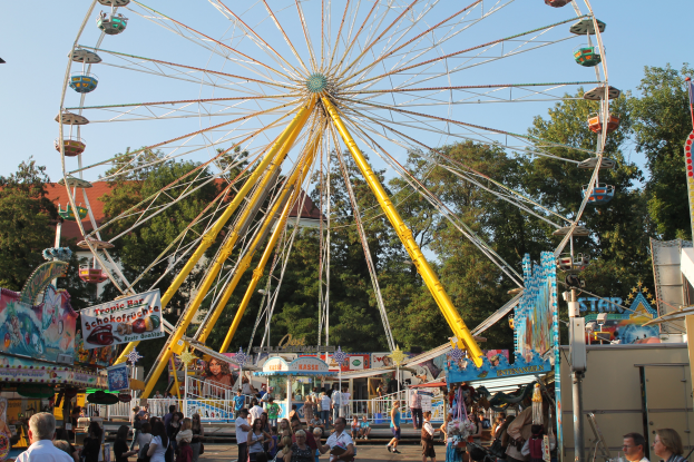Ein großes Riesenrad auf einem Rummel mit Menschen auf dem Boden, ein Stand mit Text auf der rechten Seite und Bäume mit einem klaren blauen Himmel im Hintergrund.