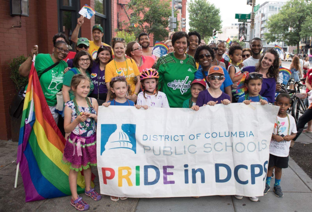 Gruppe von Menschen, die draußen stehen und ein "Bezirk Columbia Public Schools Pride in DCPS"-Schild halten, einige Kinder halten eine Fahne, eine Person auf einem Fahrrad, Bäume, Gebäude, Straßen-Elemente und ein bewölkter Himmel.